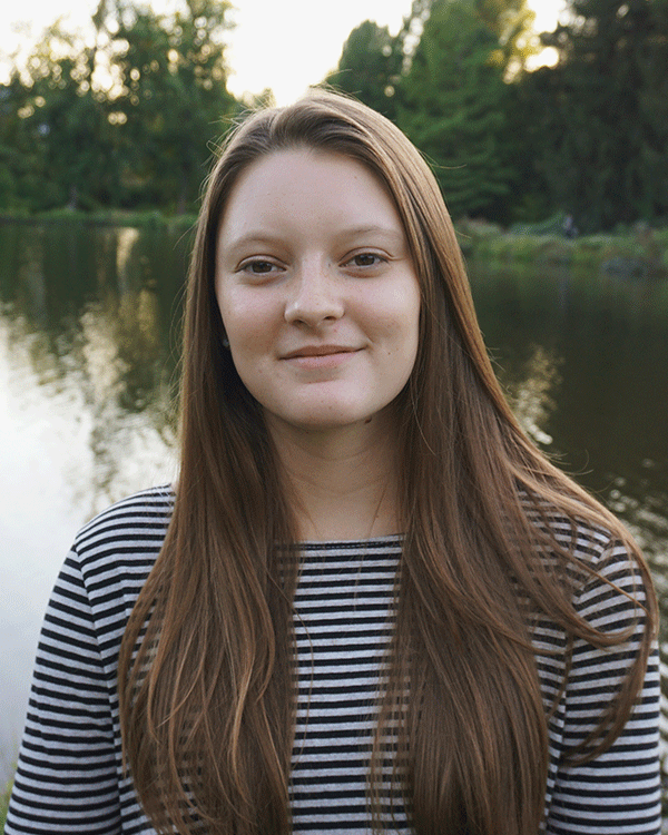 Portrait of a young woman with long brown hair, wearing a black and white striped shirt, standing by a lake with green trees in the background.