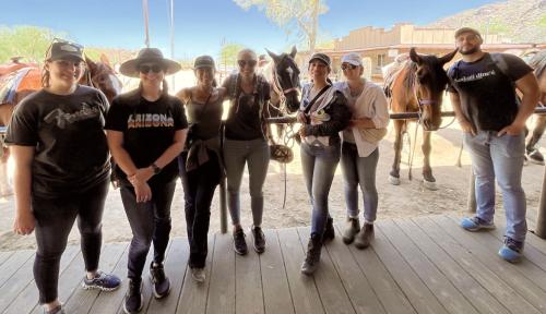 Group of seven people posing in front of saddled horses tied up on a wooden platform.