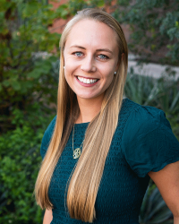 Portrait of a woman with long blonde hair in a teal dress, smiling in an outdoor setting with greenery.