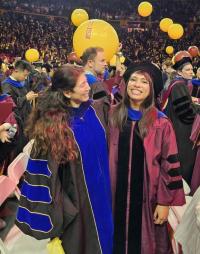 Two female graduates in black academic robes smiling, surrounded by other graduates and yellow balloons.