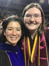 wo smiling individuals, one in academic regalia and the other in a graduation gown with stole and cords, at a graduation ceremony.