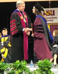 Two individuals in academic robes shaking hands on a stage at a graduation ceremony, with "Arizona State University" banner in the background.